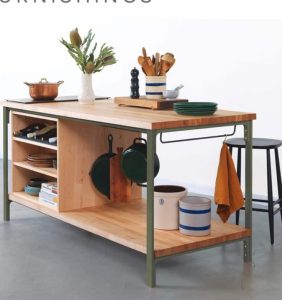 Isla de cocina con estantes abiertos y estructura de metal verde. Superficie de madera clara. Decorada con utensilios, plantas y recipientes de cocina. Ideal para almacenamiento y organización.