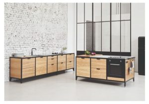 Isla de cocina minimalista con gabinetes de madera clara, encimera negra, horno empotrado y detalles metálicos, frente a pared de ladrillo blanco en ambiente moderno.