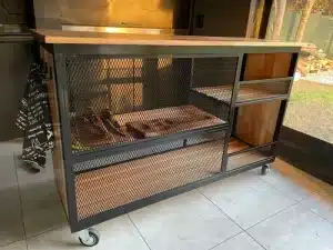 Mesa de trabajo industrial con estructura de metal negro, estantes de madera y paneles de malla. Montada sobre ruedas para facilitar su movilidad, ideal para espacios de cocina o taller.