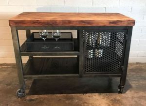 Carro de bar industrial con estructura metálica negra, encimera de madera, estantes abiertos, compartimento de rejilla y ruedas. Ideal para almacenamiento y exhibición de botellas y copas.