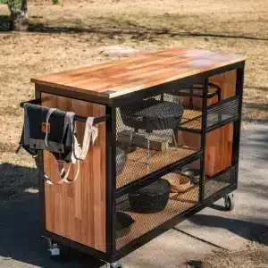 Carrito de cocina en exterior con encimera de madera, estructura de metal negro y ruedas. Incluye estantes de malla y un soporte lateral para colgar herramientas o accesorios de cocina.