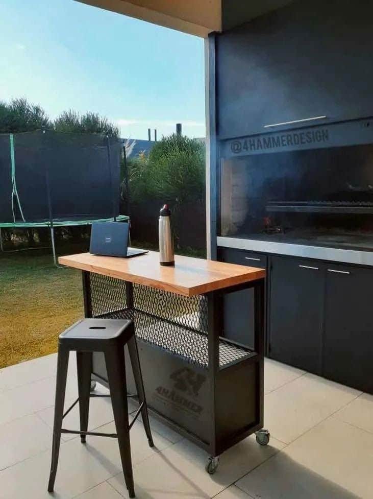 Mesa de exterior con estructura metálica negra, cubierta de madera clara y ruedas. Incluye un taburete alto a juego, ideal para cocinas o parrillas al aire libre.