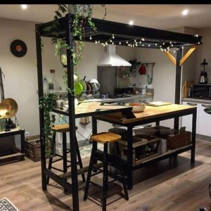 Isla de cocina de madera y metal, con taburetes altos y estantería inferior. Decorada con luces y plantas colgantes, brinda un ambiente acogedor y moderno.