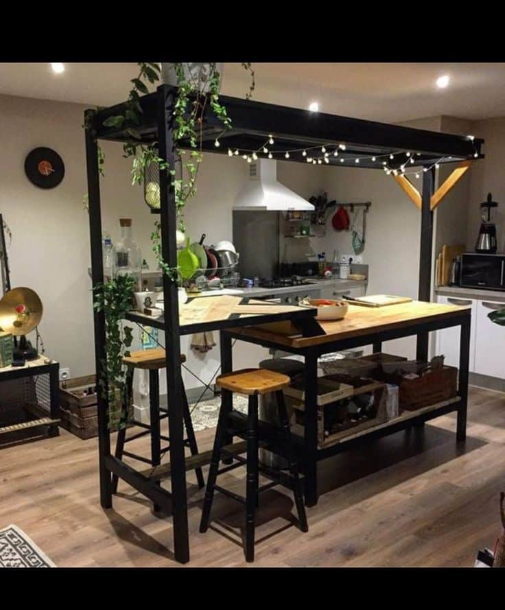 Isla de cocina de madera y metal, con taburetes altos y estantería inferior. Decorada con luces y plantas colgantes, brinda un ambiente acogedor y moderno.