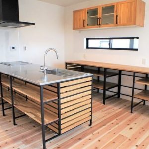 Isla de cocina moderna con estructura metálica negra y repisas de madera clara. Encimera de acero inoxidable con fregadero integrado. Estanterías abiertas y gabinetes superiores a juego.