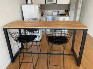 Mesa alta de madera clara con estructura metálica negra. Acompañada por cuatro sillas con diseño de rejilla metálica negra. Estilo contemporáneo en un espacio de cocina moderno.