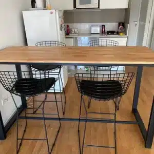 Mesa alta de madera clara con estructura metálica negra. Acompañada por cuatro sillas con diseño de rejilla metálica negra. Estilo contemporáneo en un espacio de cocina moderno.