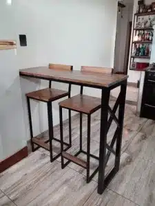 Mesa alta de diseño industrial con estructura metálica negra y superficie de madera. Incluye dos taburetes a juego con respaldo bajo, ideal para espacios reducidos.