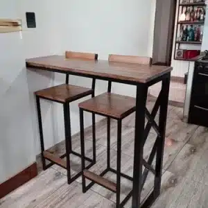 Mesa alta de diseño industrial con estructura metálica negra y superficie de madera. Incluye dos taburetes a juego con respaldo bajo, ideal para espacios reducidos.