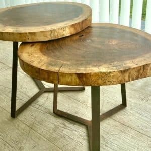 Dos mesas de centro rústicas hechas de madera maciza con bordes naturales y patas metálicas en un diseño asimétrico. La textura y los anillos de la madera destacan sobre un suelo de parqué claro.