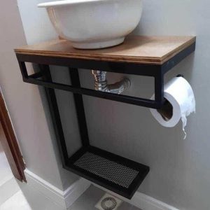 Lavabo moderno suspendido con estructura metálica negra, estante de madera y lavabo de cerámica. Incluye dispensador de jabón dorado y soporte lateral para papel higiénico.