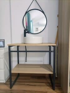 Mueble de baño moderno con estructura metálica negra y estantes de madera clara. Incluye lavabo blanco redondo en la parte superior y espejo circular con marco negro colgado encima.