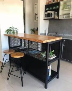 Barra alta de cocina con superficie de madera, estructura negra y estantería de malla metálica. Incluye bancos de estilo industrial con asientos de madera y patas metálicas negras.