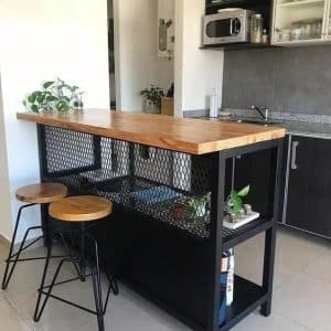 Barra alta de cocina con superficie de madera, estructura negra y estantería de malla metálica. Incluye bancos de estilo industrial con asientos de madera y patas metálicas negras.