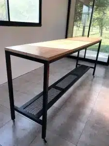 Mesa alta rectangular con estructura metálica negra y superficie de madera clara. Incluye una repisa inferior de malla. Ideal para cocina o taller. Diseño moderno y funcional.