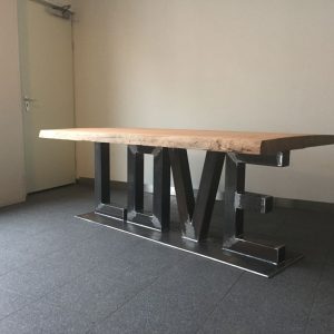 Mesa de madera con base metálica en forma de la palabra LOVE, ubicada en una sala con suelo de baldosas grises y pared blanca. Diseño moderno y artístico.