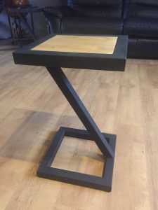 Mesa auxiliar moderna con estructura negra en forma de Z y tablero cuadrado de madera clara. Perfecta para decorar salones o salas de estar con estilo contemporáneo.