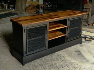 Mueble tipo aparador de estilo industrial, con estructura metálica negra, puertas de rejilla y superficie de madera pulida. Incluye estantes centrales de madera ajustables.