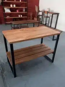 Mesa de madera con estante inferior y estructura metálica negra. Diseño robusto y moderno, ideal para cocina o sala de estar. Fondo con estantería y sillas altas.