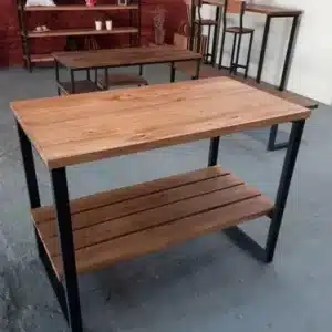 Mesa de madera con estante inferior y estructura metálica negra. Diseño robusto y moderno, ideal para cocina o sala de estar. Fondo con estantería y sillas altas.