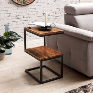 Mesa auxiliar moderna con estructura de metal negro y dos niveles de madera pulida. Ideal para espacios contemporáneos, junto a un sofá gris sobre piso de madera clara.