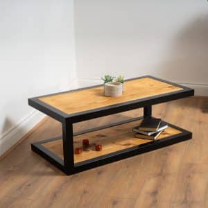 Mesa de centro moderna con estructura metálica negra y superficie de madera clara. Cuenta con dos niveles, siendo el inferior usado para almacenar libros y decoraciones. Ideal para sala de estar.