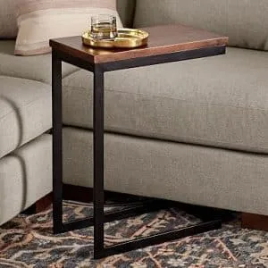 Mesa auxiliar contemporánea en forma de C con base de metal negra y superficie de madera oscura, ubicada junto a un sofá gris. Ideal para pequeños espacios.