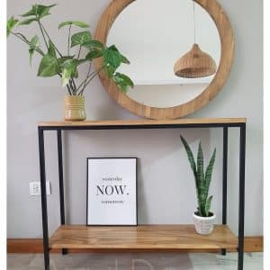 Mesa consola de madera clara con patas negras de metal. Incluye estante inferior y está adornada con plantas. Encima cuelga un espejo redondo con marco de madera a juego.