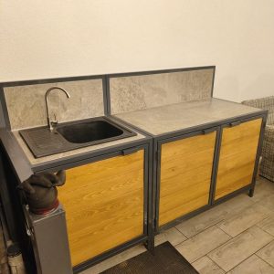 Gabinete de cocina con puertas de madera y estructura metálica; incluye un fregadero negro y encimeras de piedra. Diseño industrial y rústico, ideal para espacios modernos.