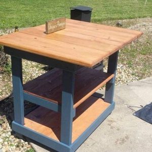 Mesa de madera en exterior, estructura azul con dos estantes inferiores y superficie superior amplia, ideal para jardines o patios.