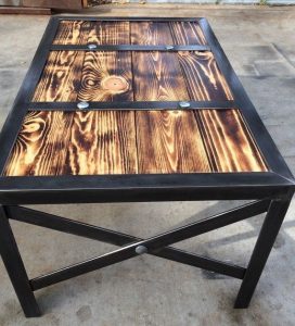 Mesa de centro cuadriculada, con tablero de madera quemada y vetas visibles, enmarcada en metal negro, y patas cruzadas con remaches visibles. Estilo rústico e industrial.