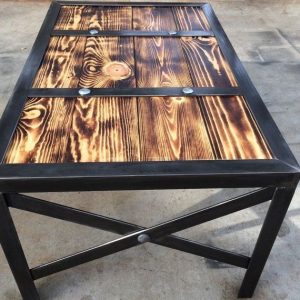 Mesa de centro cuadriculada, con tablero de madera quemada y vetas visibles, enmarcada en metal negro, y patas cruzadas con remaches visibles. Estilo rústico e industrial.