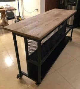 Mesa de cocina industrial con superficie de madera rústica y estructura metálica negra. Cuenta con ruedas y estantes de rejilla para almacenamiento. Ideal para espacios modernos.