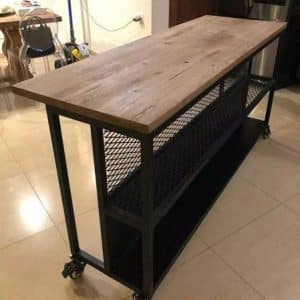 Mesa de cocina industrial con superficie de madera rústica y estructura metálica negra. Cuenta con ruedas y estantes de rejilla para almacenamiento. Ideal para espacios modernos.