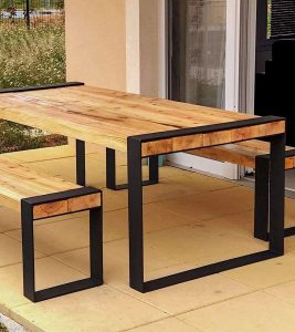 Mesa de madera con estructura metálica negra minimalista. Tiene un acabado natural y está acompañada por un banco a juego, ideal para exteriores.