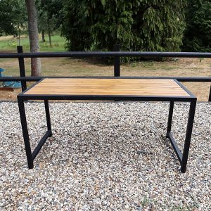 Mesa de exterior con superficie de madera y estructura de metal negro, ubicada sobre un suelo de grava en un entorno natural.