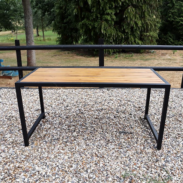 Mesa de exterior con superficie de madera y estructura de metal negro, ubicada sobre un suelo de grava en un entorno natural.