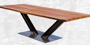 Mesa de comedor con cubierta de madera maciza, forma rectangular. Base en forma de V invertida de metal oscuro sobre plataforma rectangular del mismo material. Diseño moderno.