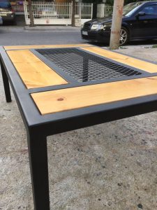 Mesa de exterior con estructura de metal negro, tablones de madera clara y rejilla central metálica. Diseño moderno y resistente para jardines o terrazas.