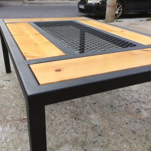 Mesa de exterior con estructura de metal negro, tablones de madera clara y rejilla central metálica. Diseño moderno y resistente para jardines o terrazas.