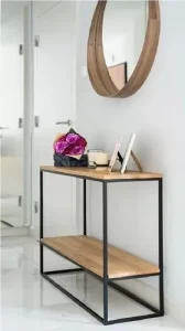 Mesa de entrada de madera clara con estructura de metal negro y dos niveles. Decorada con flores, portarretratos y una vela, situada junto a una pared blanca y un espejo redondo.
