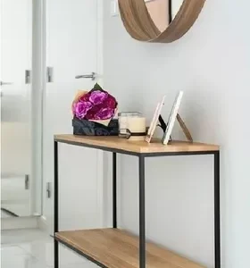 Mesa de entrada de madera clara con estructura de metal negro y dos niveles. Decorada con flores, portarretratos y una vela, situada junto a una pared blanca y un espejo redondo.