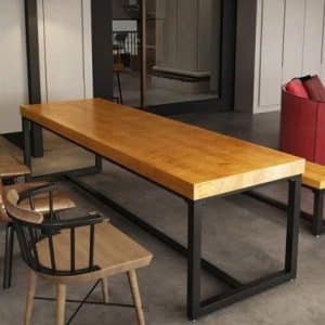 Mesa de comedor rectangular de madera con tapa gruesa y base de metal negro. Diseño industrial y moderno. Rodeada de sillas de madera con respaldo de barras y estructura metálica.
