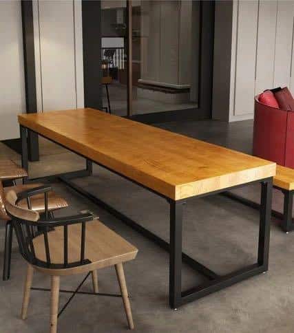 Mesa de comedor rectangular de madera con tapa gruesa y base de metal negro. Diseño industrial y moderno. Rodeada de sillas de madera con respaldo de barras y estructura metálica.