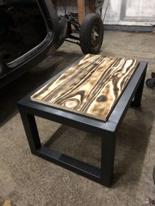 Mesa de centro con base de metal negro y tapa de madera con vetas onduladas y efecto quemado, situada sobre piso de concreto en un garaje.