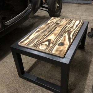 Mesa de centro con base de metal negro y tapa de madera con vetas onduladas y efecto quemado, situada sobre piso de concreto en un garaje.