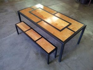 Mesa y banco con estructura negra de metal y paneles de madera clara con diseño geométrico rectangular, ubicados en un piso de concreto.