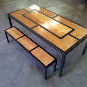 Mesa y banco con estructura negra de metal y paneles de madera clara con diseño geométrico rectangular, ubicados en un piso de concreto.