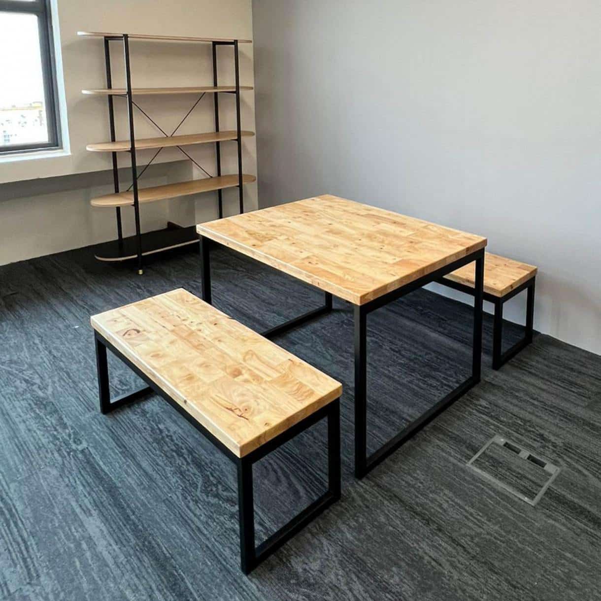 Juego de comedor moderno con mesa cuadrada de madera clara y metal negro, acompañado de dos bancos a juego. Al fondo, estantería esquinera con varias repisas. Estilo industrial.