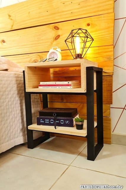 Mesa de noche de madera y metal con dos estantes. Incluye decoración con libros, cajas y una lámpara geométrica. Diseño moderno y funcional para dormitorios.